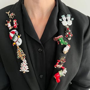 Puede incluir: Una colección de broches navideños con diseños variados, incluyendo muñecos de nieve, árboles de Navidad, renos y figuras de Papá Noel. Los broches son coloridos y detallados, con acentos rojos, verdes, blancos y dorados, y se exhiben en una blazer negra.