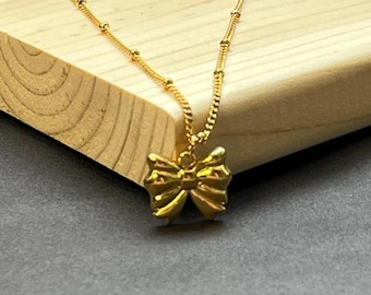 Puede incluir: Un collar dorado con un colgante de lazo. El lazo es detallado y brillante. La cadena del collar también es dorada y tiene un diseño delicado. El collar se muestra sobre una superficie de madera.