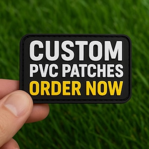 Pode incluir: Um patch retangular preto de PVC com texto branco e amarelo. O texto diz "CUSTOM PVC PATCHES ORDER NOW". O patch é mantido contra um fundo de grama verde.