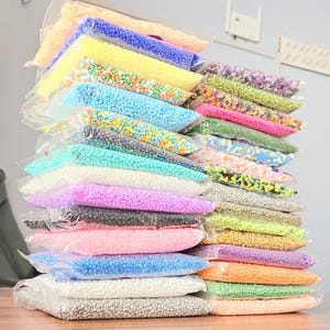 May include: A stack of clear plastic bags filled with colorful beads. The beads are in various colors, including blue, yellow, pink, green, and purple. The bags are stacked on top of each other, creating a visually appealing display.