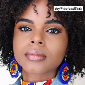 May include: A woman with dark curly hair wearing large blue beaded earrings and a matching beaded necklace. The earrings and necklace feature colorful patterns of orange, yellow, and red. The text "etsy/WaistBeadDeals" is visible.