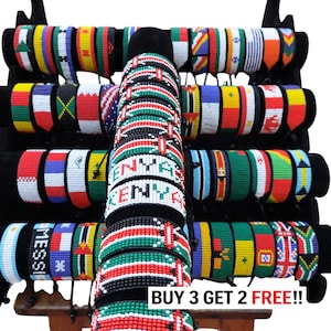 May include: A collection of beaded bracelets displayed on a black stand, featuring various national flag patterns and designs. Several bracelets display the word KENYA in white beads, while one bracelet features the text MESSI. Text reads BUY 3 GET 2 FREE!!