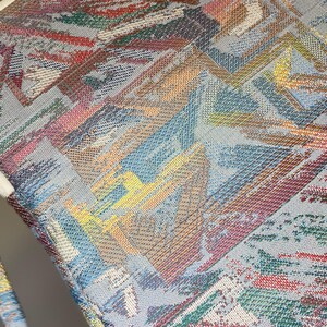 May include: Close-up of a woven textile with a geometric pattern. The design features a mix of colors including blue, red, green, yellow, and brown. The fabric has a textured appearance, with a light blue background.