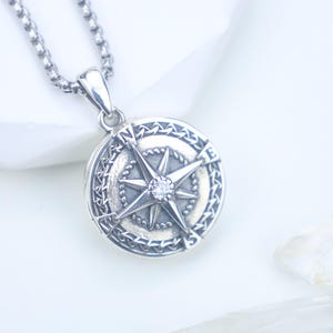 May include: A silver-toned compass pendant necklace. The pendant features a detailed compass rose design with a central gemstone. The necklace has a chain with a textured, rounded link design. The pendant is round and has a detailed border.