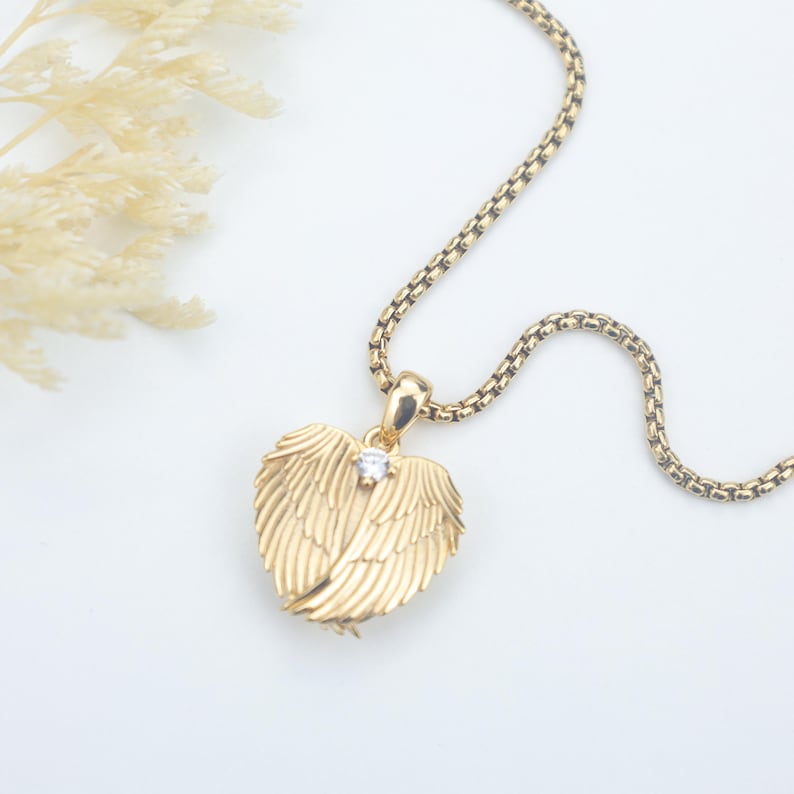 Puede incluir: Un collar dorado con un colgante en forma de alas de &aacute;ngel. El colgante tiene una peque&ntilde;a gema transparente en el centro. La cadena es de color dorado a juego y tiene un dise&ntilde;o texturizado y tejido. El collar se muestra sobre una superficie blanca.