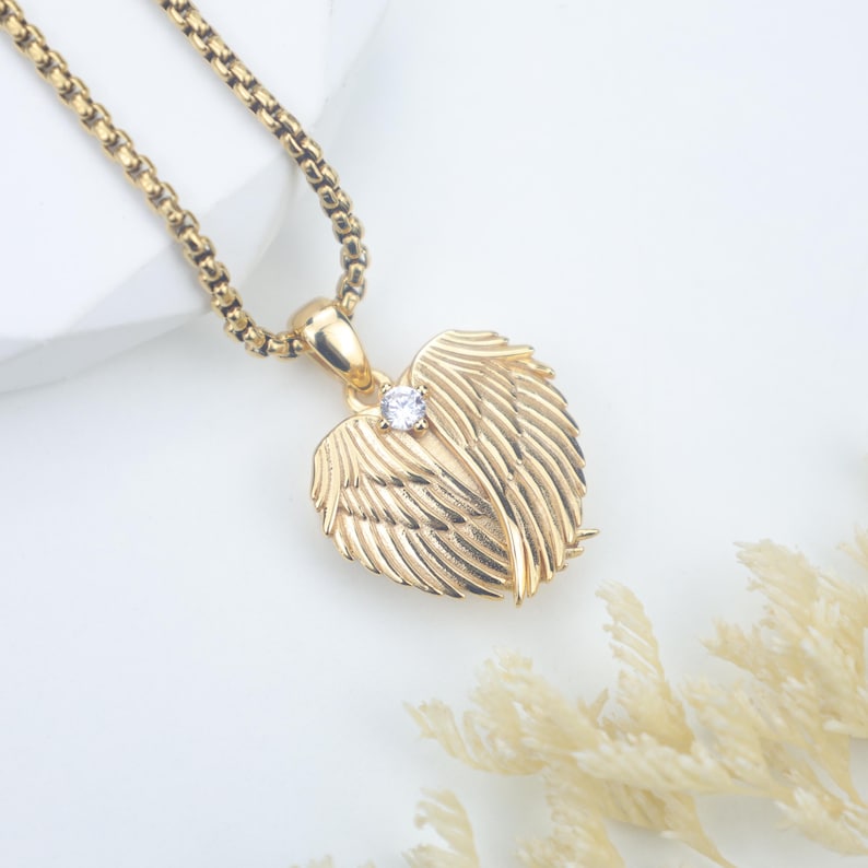 May include: Gold-toned necklace featuring a pendant shaped like angel wings. The pendant has a small, clear gemstone at its center. The chain is a box chain style. The necklace is displayed on a white surface.