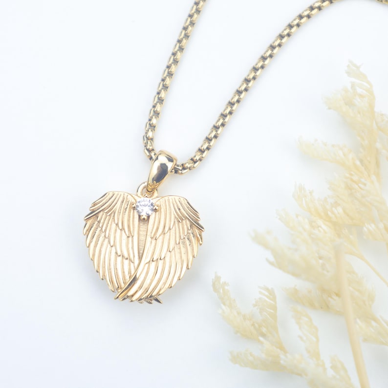 Puede incluir: Un collar con colgante dorado con un dise&ntilde;o en forma de coraz&oacute;n con alas de &aacute;ngel. Una peque&ntilde;a gema transparente est&aacute; engastada en el centro del colgante. La cadena del collar tambi&eacute;n es dorada y tiene un patr&oacute;n texturizado y enlazado.