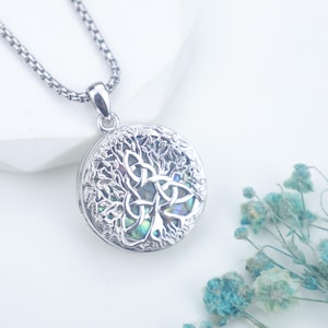 Op de afbeelding: Een zilverkleurige ketting met een ronde hanger. De hanger heeft een Keltisch levensboomontwerp met gedetailleerde uitgesneden patronen en iriserende accenten. De ketting is zilverkleurig en heeft een getextureerd ontwerp.