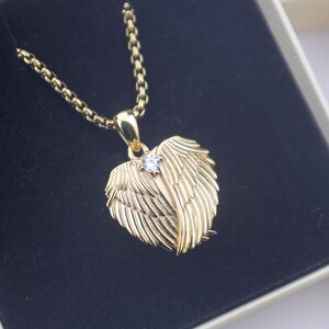 Puede incluir: Un collar dorado con un colgante en forma de coraz&oacute;n. El colgante presenta un dise&ntilde;o de alas con una peque&ntilde;a gema transparente en el centro. El collar se muestra en una caja negra.