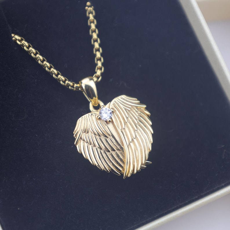 Puede incluir: Un collar dorado con un colgante en forma de coraz&oacute;n. El colgante presenta un dise&ntilde;o de alas con una peque&ntilde;a gema transparente en el centro. El collar se muestra en una caja negra.