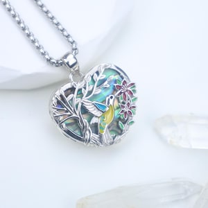 Puede incluir: Collar con colgante en forma de corazón plateado con diseño de colibrí. El colgante presenta un fondo colorido de concha de abulón, un colibrí con detalles amarillos y azules y flores rojas. La cadena del collar también es plateada.