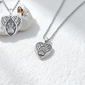 May include: Silver heart pendant necklace with a paw print design. The pendant has angel wings and is decorated with clear crystals. A small heart-shaped charm with a dog photo is also visible.