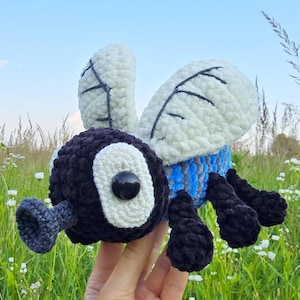 Puede incluir: Un juguete de mosca hecho a mano con ganchillo, con un cuerpo negro, alas blancas con costuras negras y un abdomen a rayas azules y negras. La mosca tiene ojos blancos grandes con pupilas negras. Se sostiene frente a un campo de hierba.