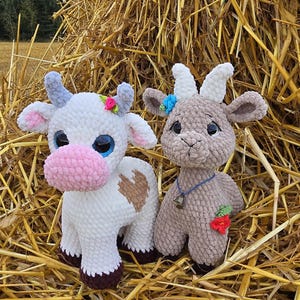 May include: Two handmade crochet stuffed animals: a white cow with a pink nose and a brown goat. The cow has blue horns and a floral crown. The goat has white horns and a blue necklace with a bell and a red strawberry.