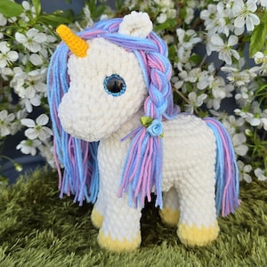 May include: A white crocheted unicorn with a yellow horn, blue and pink mane, and blue eyes. The unicorn has yellow hooves and a small blue flower. It is sitting on green faux grass, with white flowers in the background.