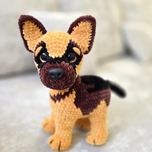 Puede incluir: Un perro pastor alemán de ganchillo hecho a mano. El peluche presenta una combinación de colores marrón y beige, con nariz y ojos negros. El perro está sentado, con las orejas levantadas. El juguete está hecho de hilo suave y texturizado.