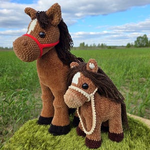 May include: Two plush, brown crochet horses stand on a green surface against a backdrop of a field and blue sky. The larger horse has a red halter, while the smaller one has a cream-colored halter. Both have dark brown manes and black hooves.