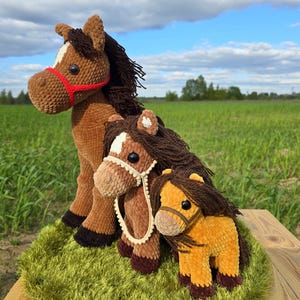 Puede incluir: Tres figuras de caballos de ganchillo de diferentes tamaños, marrones, beige y amarillos, con crines y colas marrones oscuras. El caballo más grande tiene un cabestro rojo, los otros beige. Están sobre una superficie verde texturizada. Fondo verde y cielo azul.