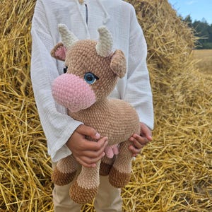 May include: A crochet cow plush toy in brown, beige, and pink. The cow has blue eyes, white horns, and a pink nose. The toy is held against a backdrop of hay bales.