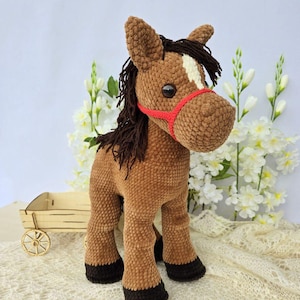 May include: A handmade brown crochet horse with a red halter. The horse has a white stripe on its face and dark brown mane and tail. The horse has black hooves and large black eyes. The horse is a stuffed animal.
