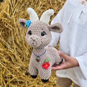 Op de afbeelding: Een handgemaakte gehaakte geit knuffel in bruin, wit en zwart. De geit heeft witte hoorns, een blauwe bloem en een rode bloem met groene bladeren. Het draagt een blauwe ketting met een bel. De knuffel wordt voor een hooibaal gehouden.