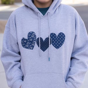 May include: A pale grey hoodie with a front pocket and a drawstring hood. The hoodie has three blue heart appliqués with different patterns: floral, polka dots, and floral. A teal shirt is visible beneath the hoodie.