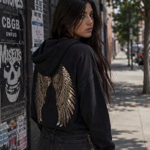 May include: A black hooded sweatshirt featuring a large, metallic gold sequined wing design on the back. The hoodie is paired with black denim shorts that have a silver chain attached to the side, creating a cohesive dark-toned outfit.