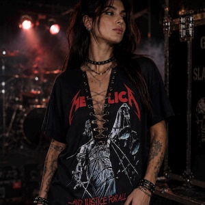Op de afbeelding: Een zwart T-shirt met korte mouwen en een Metallica-afbeelding met de tekst "METALLICA" en "...AND JUSTICE FOR ALL" in het rood. Het shirt heeft een diepe V-hals met een veterdetail door oogjes met gekruiste veters in het midden.
