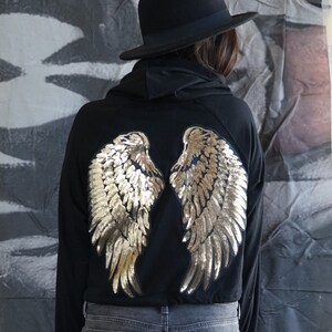 May include: Black hoodie with gold sequined angel wings on the back. The wings are detailed with individual sequins, creating a shimmering effect. The hoodie is paired with gray jeans and a black hat.