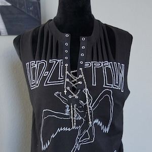 May include: Black sleeveless shirt with the Led Zeppelin logo and a graphic of a winged figure, with the text "UNITED STATES OF AMERICA 1977". The shirt has a lace-up neckline with silver grommets and a silver chain.