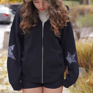 May include: A black zip-up hoodie with a star-shaped design on each sleeve. The hoodie has a relaxed fit and a drawstring hood.