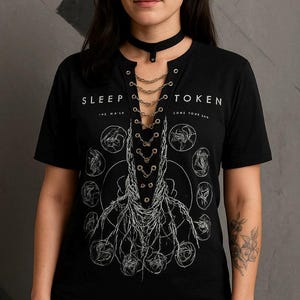 May include: Black t-shirt with the text "SLEEP TOKEN" and a graphic design. The shirt has a V-neck with chain detailing and is paired with a black choker. The design features a root-like illustration.
