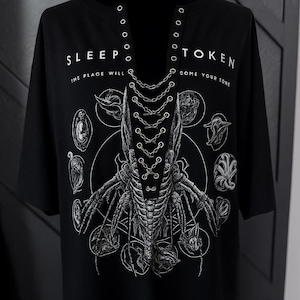 May include: Black t-shirt with a unique graphic design featuring a central skeletal figure with intricate details and surrounding circular illustrations. The text "SLEEP TOKEN" and "COME YOUR TONE" are printed on the shirt.