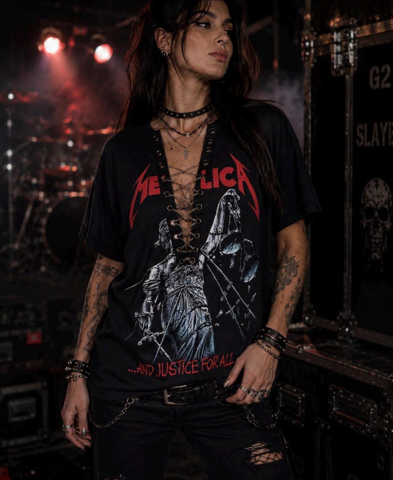Op de afbeelding: Een zwart T-shirt met korte mouwen en een Metallica-afbeelding met de tekst "METALLICA" en "...AND JUSTICE FOR ALL" in het rood. Het shirt heeft een diepe V-hals met een veterdetail door oogjes met gekruiste veters in het midden.