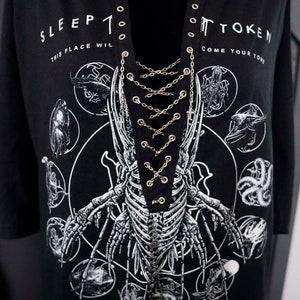 May include: Black long-sleeve shirt with a graphic of a skeleton and the text "SLEEP TOKEN". The shirt has a lace-up neckline with silver chain detailing. The design includes various circular illustrations.