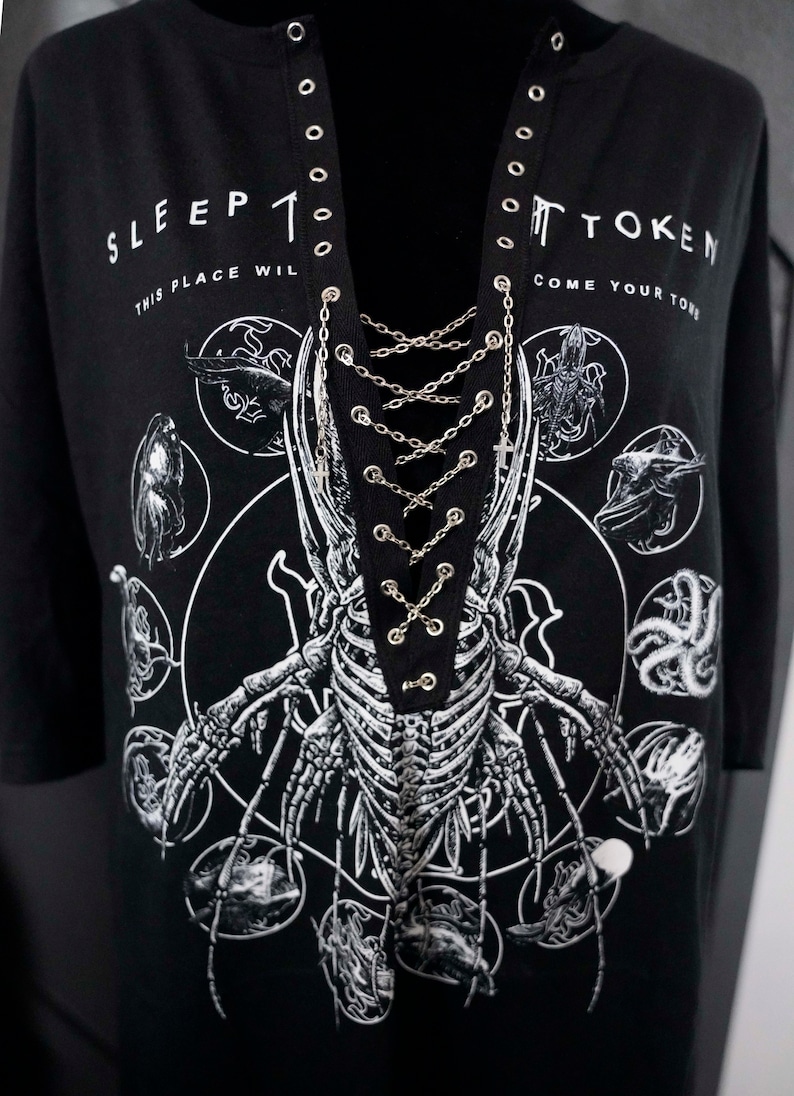 May include: Black long-sleeve shirt with a graphic of a skeleton and the text "SLEEP TOKEN". The shirt has a lace-up neckline with silver chain detailing. The design includes various circular illustrations.