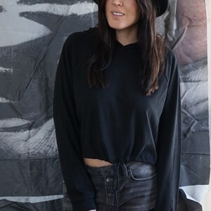 May include: A black hoodie with a drawstring waist and long sleeves, paired with denim shorts. The model is wearing a black hat. The background features a black and white abstract design.