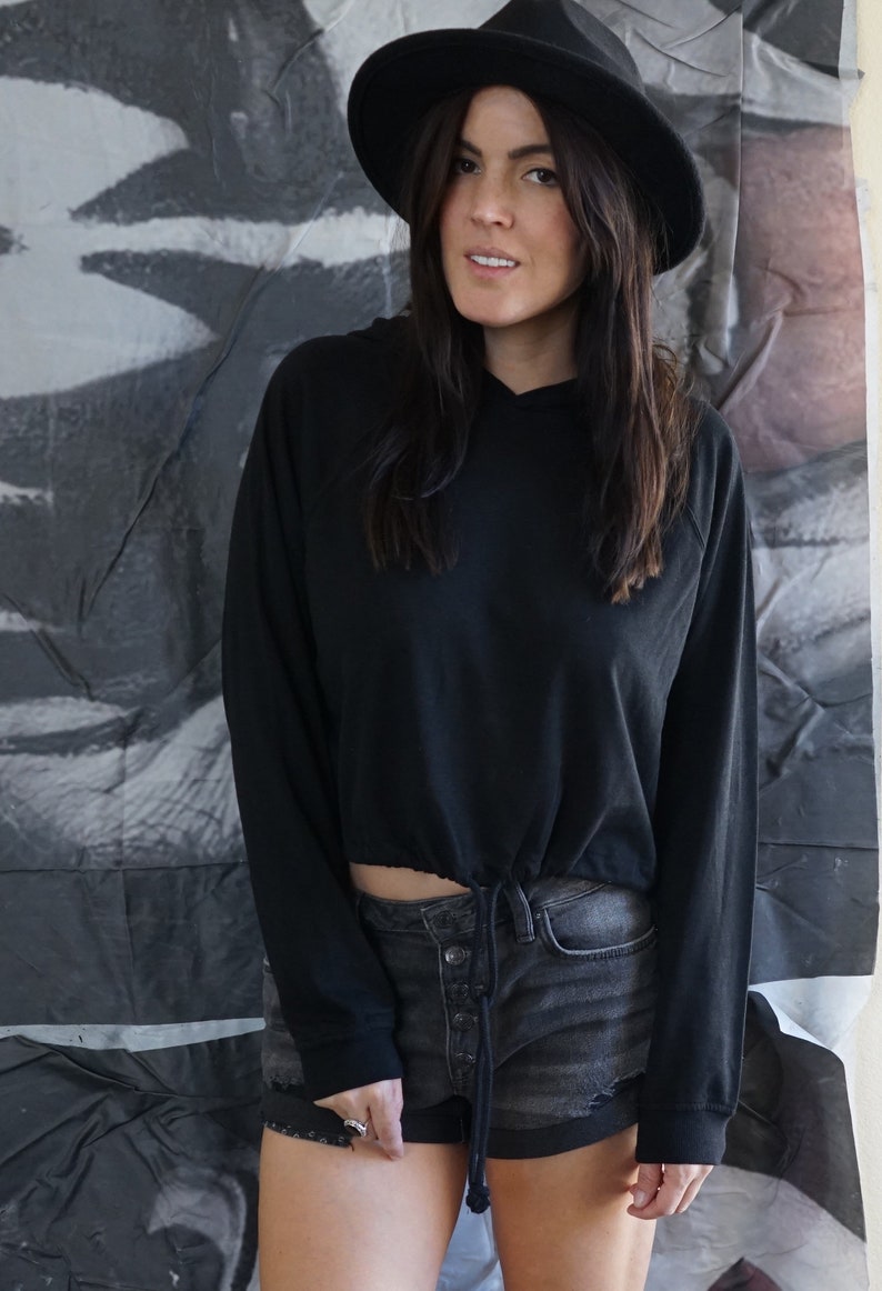 May include: A black hoodie with a drawstring waist and long sleeves, paired with denim shorts. The model is wearing a black hat. The background features a black and white abstract design.
