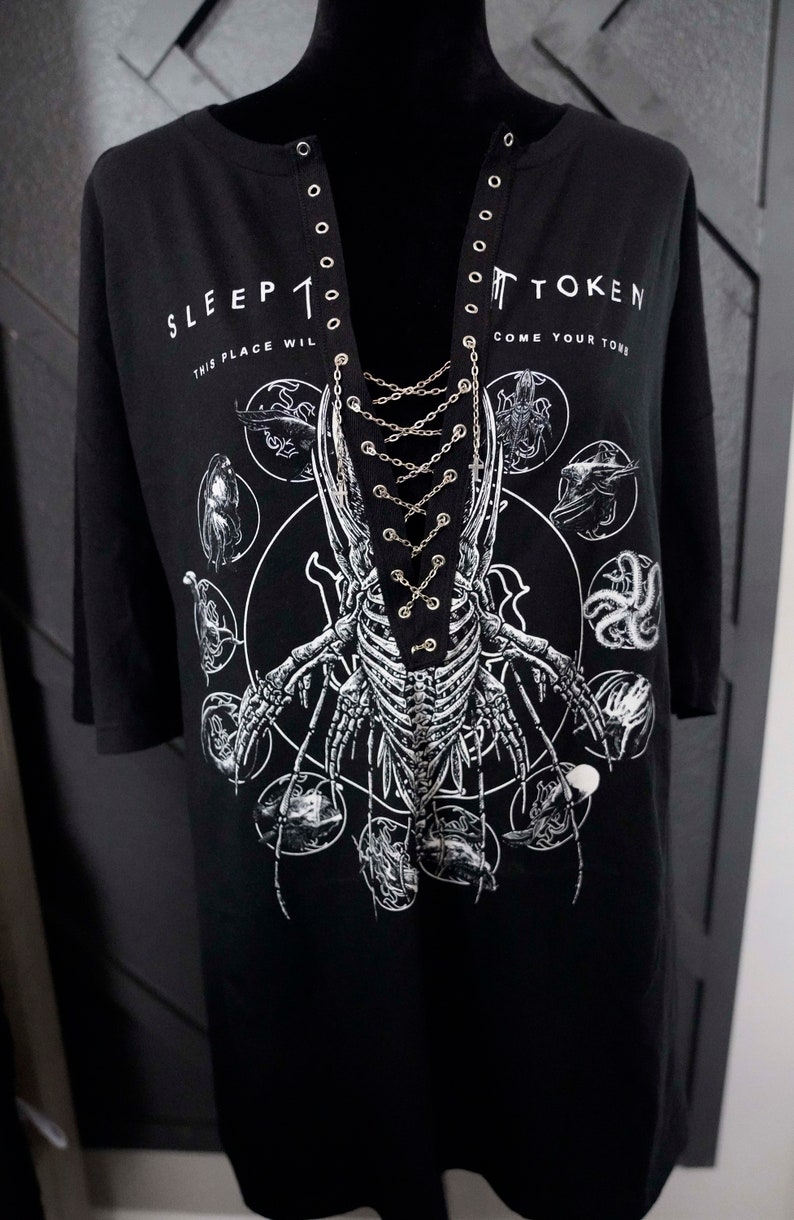 May include: Black t-shirt with a unique design featuring a skeletal insect-like figure and the text "SLEEP TOKEN" and "COME YOUR TOME". The shirt has a lace-up neckline with metal eyelets and chain detailing.