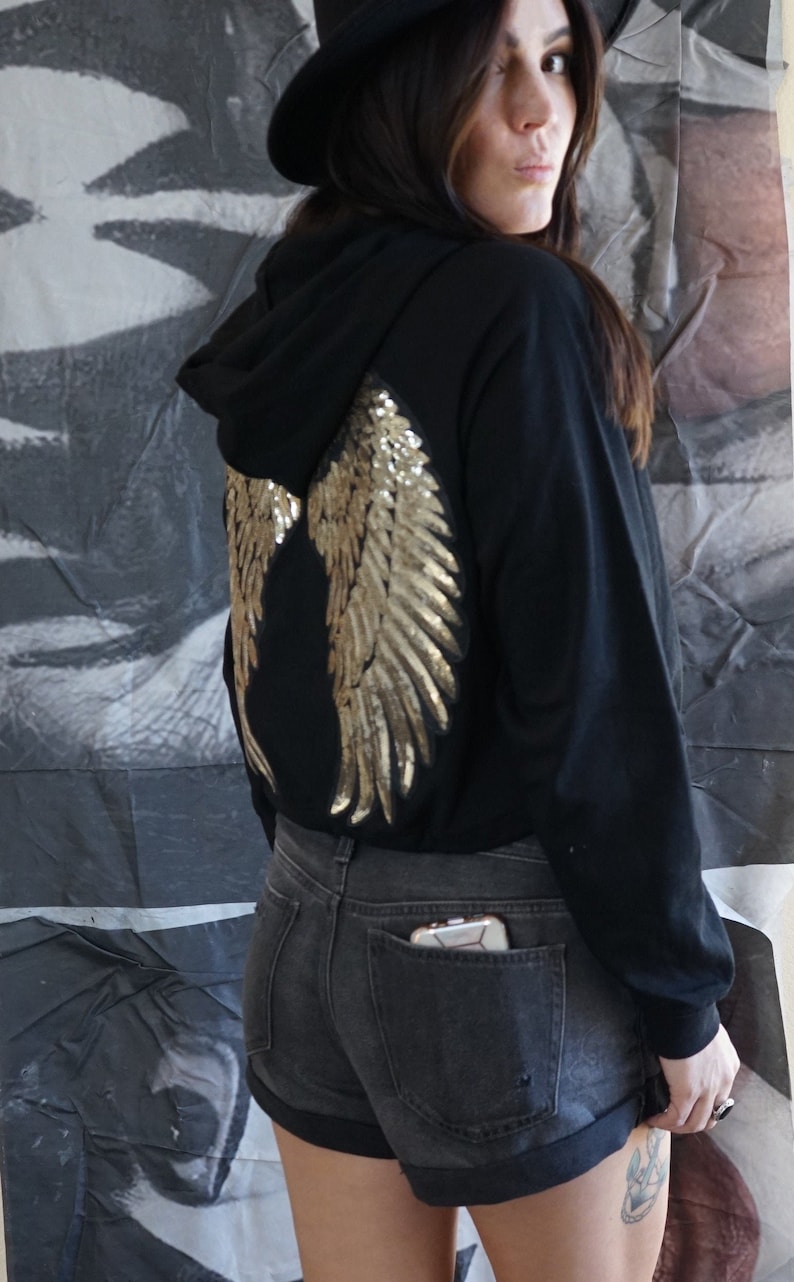 May include: Black hoodie with gold sequin angel wings on the back. The model is wearing black denim shorts and a black hat. The hoodie has a hood and long sleeves.