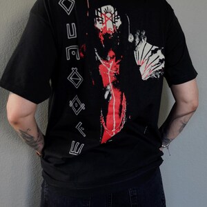 Puede incluir: Camiseta negra con un estampado gr&aacute;fico de una persona con la cara roja y marcas blancas y rojas. El texto "ETERNAL" est&aacute; impreso verticalmente en el lado izquierdo de la camiseta.