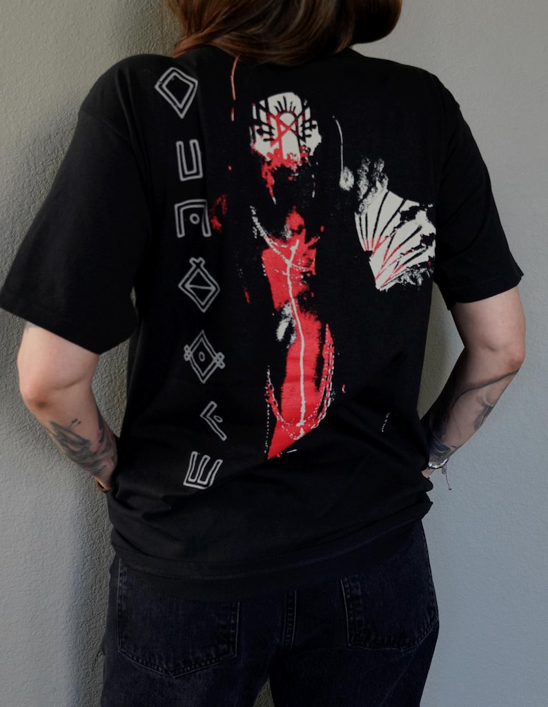 Puede incluir: Camiseta negra con un estampado gr&aacute;fico de una persona con la cara roja y marcas blancas y rojas. El texto "ETERNAL" est&aacute; impreso verticalmente en el lado izquierdo de la camiseta.