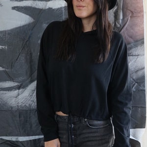 May include: A black hooded sweatshirt with a drawstring waist and long sleeves. The model is wearing black denim shorts and a black hat. The background is a gray and white abstract design.