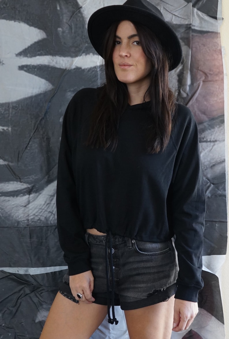 May include: A black hooded sweatshirt with a drawstring waist and long sleeves. The model is wearing black denim shorts and a black hat. The background is a gray and white abstract design.