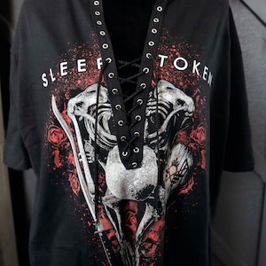 Op de afbeelding: Zwart T-shirt met een grafische print van een skelet met een zwaard en rozen. Het shirt heeft een kanten halslijn en de tekst "SLEEP FOR TOKEN" staat op de voorkant.