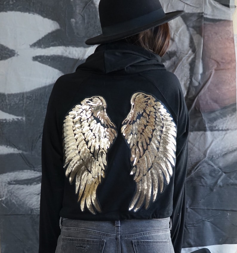 May include: Black hoodie with gold sequined angel wings on the back. The wings are detailed with individual sequins, creating a shimmering effect. The hoodie is paired with gray jeans and a black hat.