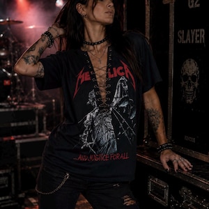 Op de afbeelding: Een zwart t-shirt met korte mouwen, voorzien van een rood Metallica-logo en een afbeelding van Vrouwe Justitia met een weegschaal. Het shirt heeft een diepe V-hals met een vetersluiting door metalen oogjes en de tekst ...AND JUSTICE FOR ALL.