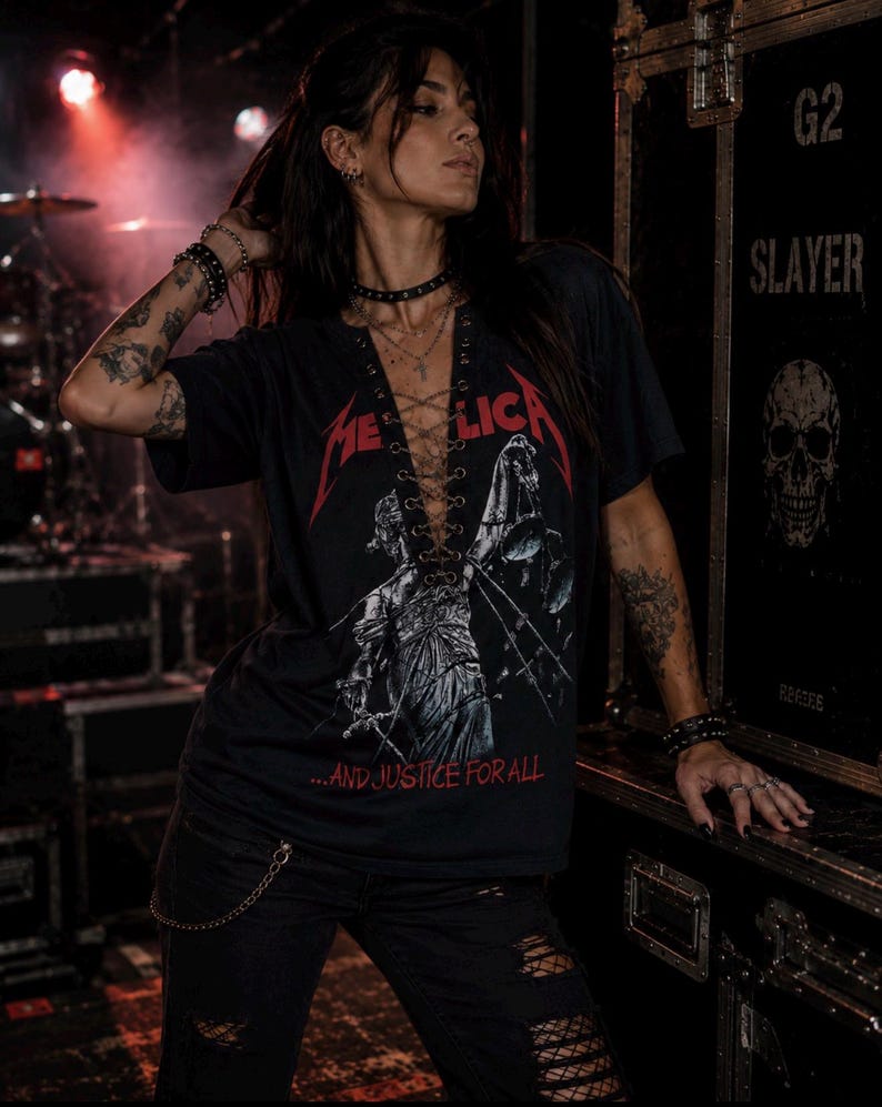 Op de afbeelding: Een zwart t-shirt met korte mouwen, voorzien van een rood Metallica-logo en een afbeelding van Vrouwe Justitia met een weegschaal. Het shirt heeft een diepe V-hals met een vetersluiting door metalen oogjes en de tekst ...AND JUSTICE FOR ALL.