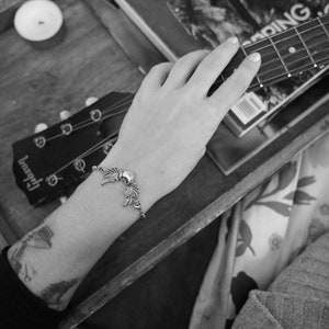 May include: A black and white photo of a person's hand holding a guitar. The guitar is a Gibson and has a silver moon and star charm bracelet on the wrist.