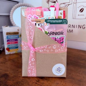 May include: A brown paper gift envelope tied with a pink ribbon featuring a white heart pattern. The envelope contains a Garnier biodegradable face mask, a pink pen, and various stickers, with a small circular sticker labelled Mo's Design Studio.